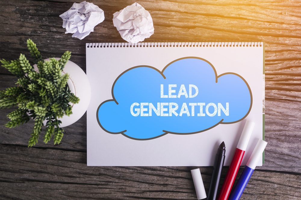 The words "Lead Generation" are written in the drawing of a cloud on a notepad. Beside it is a plant and some crumpled pieces of paper. Various marker colours lie beside the drawing.